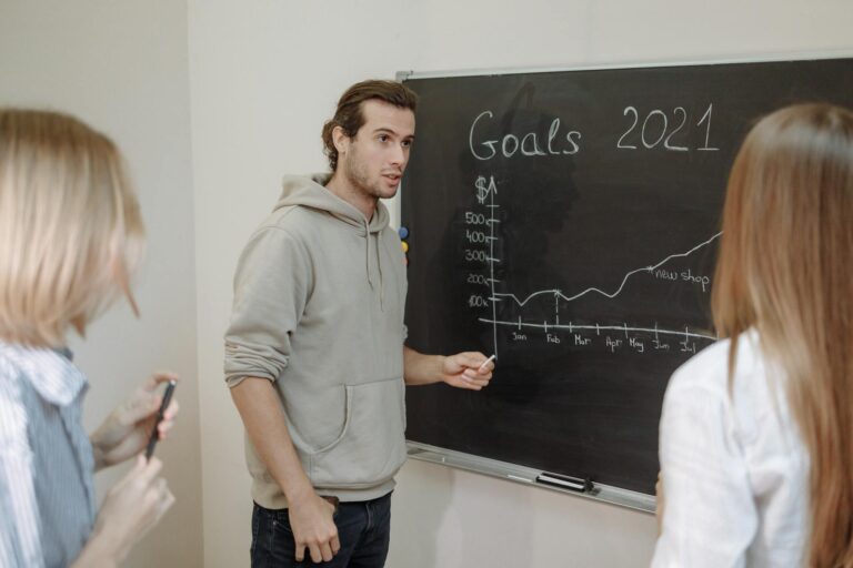 Three professionals engaged in a business discussion about goals written on a chalkboard in an office.