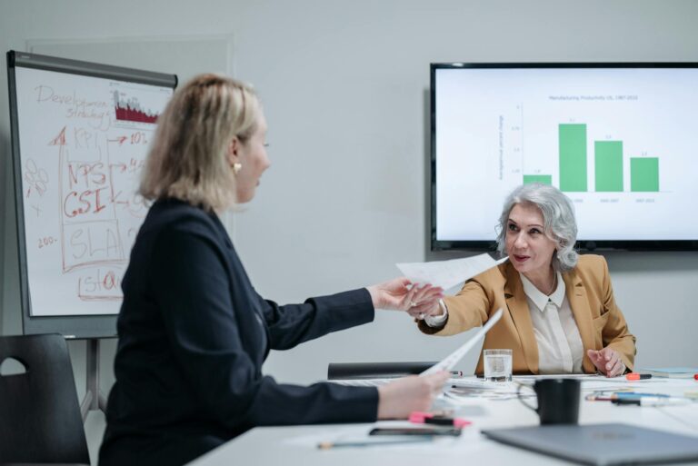Two businesswomen exchanging documents in a modern office setting with presentation screens.